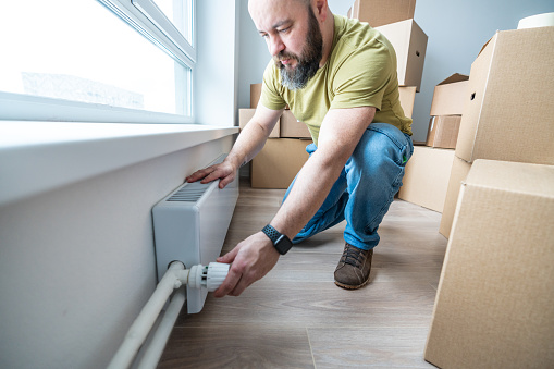 A man checking regulator of a heating radiator in the apartment full of cardboard boxes