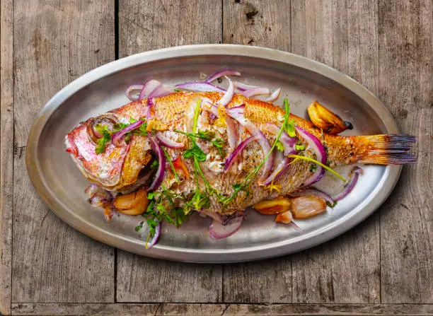 Baked Red Sea Bream, with garlics, red onion and parsley, plated on a metallic tray, over a rustic wooden table.