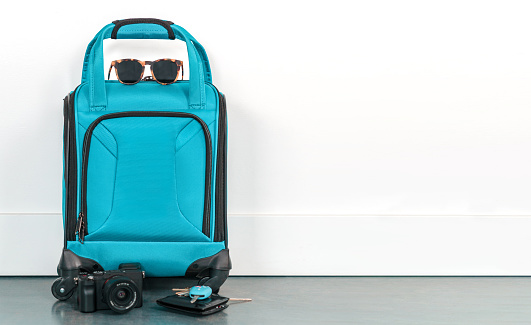 Turquoise blue travel bag with a compact camera, black wallet, car keys, and tortoise shell sunglasses in front of a white wall. Getting ready to leave for a vacation getaway. Blank wall with copy space.