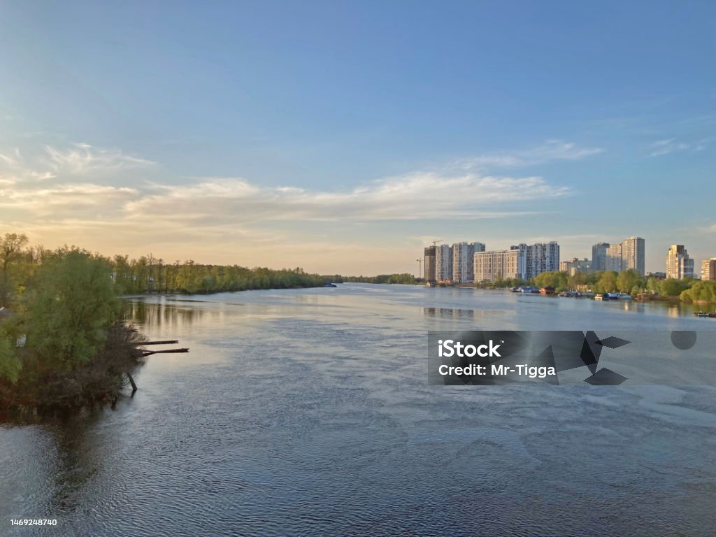 View of the deep river with the buildings in the background. Beautiful panoramic view of the city of Kyiv Dneper River at sunset on dark blue water with glowing reflected rays of the sun, copy-space Beautiful panoramic view of the city of Kyiv Dneper River at sunset on dark blue water with glowing reflected rays of the sun, copy-space. View of the deep river with the buildings in the background. Architecture Stock Photo