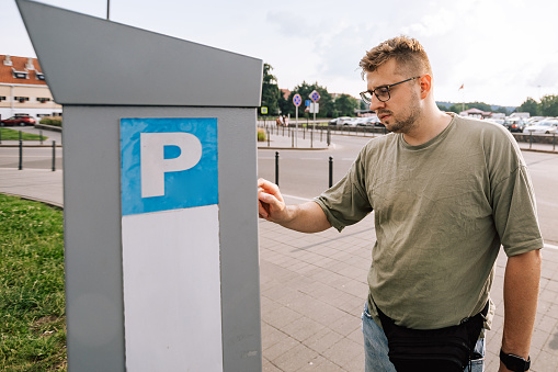 Person is using parking terminal for paying the station. Male portrait of man wear t-shirt and eyeglasses outdoors. Lifestyle photography concept.