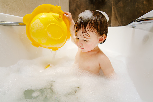 A happy child is playing with toys while sitting in a white homemade bathtub. Little toddler baby boy bathes in a bubble bath. Kid aged one year six months