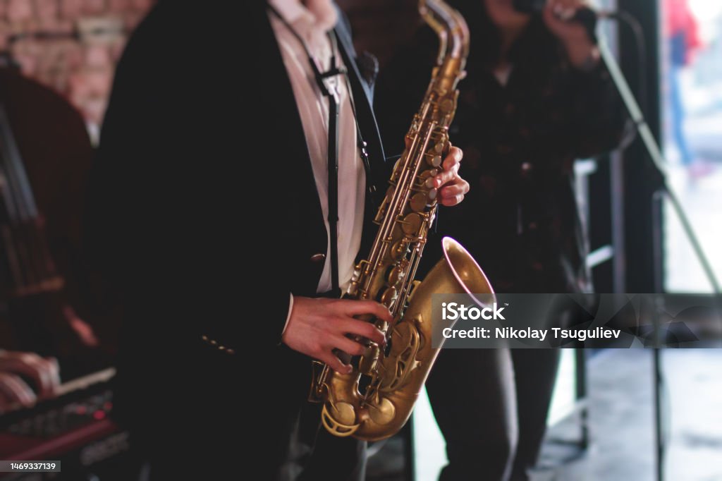 Concert view of saxophonist, a saxophone sax player with vocalist and musical band during jazz orchestra show performing music on a stage in the scene lights Concert view of saxophonist on a stage, a saxophone sax player with vocalist and musical band during jazz orchestra show performing music on a stage in the lights Adult Stock Photo
