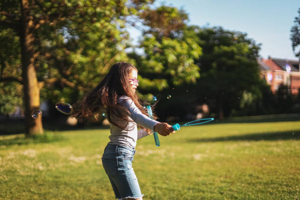 Portrait of a caucasian girl blows soap bubbles circling in the park on a playground meadow, Portrait of a beautiful caucasian girl with long flowing brown hair, a gray long-sleeved t-shirt and glasses blows soap bubbles circling in the park on a playground meadow, close-up side view. The concept of PARKS and REC, happy childhood, children's picnic, holidays, fabulous childhood, outdoor recreation, playgrounds, outdoors, soap bubbles. funny camping signs pictures stock pictures, royalty-free photos & images