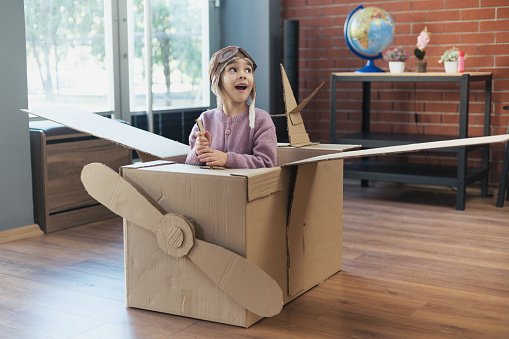 Little child girl having fun playing with the plane from cardboard at home