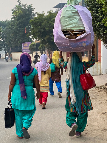 Delhi, India - November 3, 2022: Stock photo showing rear view of pile of flattened cardboard boxes being transported on the head of an unrecognisable Indian woman walking with a group of women wearing Salwar Kameez traditional clothing, on road side.