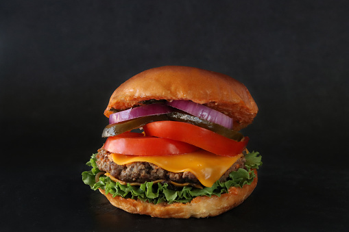 Stock photo showing a close-up view of the components of a home-made gourmet cheeseburger with beef burger in burger bun consisting of hamburger patty, melting processed cheese slices, lettuce leaves, sliced tomato, pickles and red onion slices, against a black background.