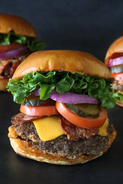 istock Close-up image of three homemade hamburger bread buns, cheeseburger, chicken, bacon cheese burgers, meat patties, bacon rashers, melting cheese slices, lettuce leaves, sliced tomato, pickles and red onion slices, black background 1469372956