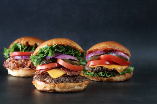 Close-up image of three homemade hamburger bread buns, cheeseburger, chicken, bacon cheese burgers, meat patties, bacon rashers, melting cheese slices, lettuce leaves, sliced tomato, pickles and red onion slices, black background, copy space