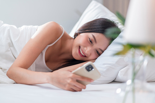 Asian adult woman in pyjamas casual relax lying on the white soft bed morning light wake up hand surfing using smartphone for shopping online text chatting or contact friends on social media. smiling cheerful facial expression