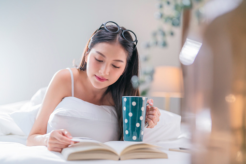 Beautiful woman lying on the bed and reading a book at home,woman lying on her bed and reading an analog book. Spending time at home during weekend. Simple pleasures in her bedroom at home