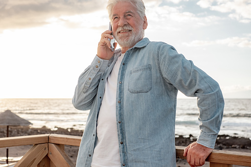 Portrait of beautiful senior bearded man in casual shirt standing outdoors at beach in sunset light using mobile phone. Smiling old man talking on smartphone