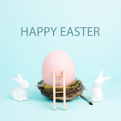 Easter bunny or rabbit sitting next to a huge pink colored egg in a birds nest, paint brush and ladder, spring holiday