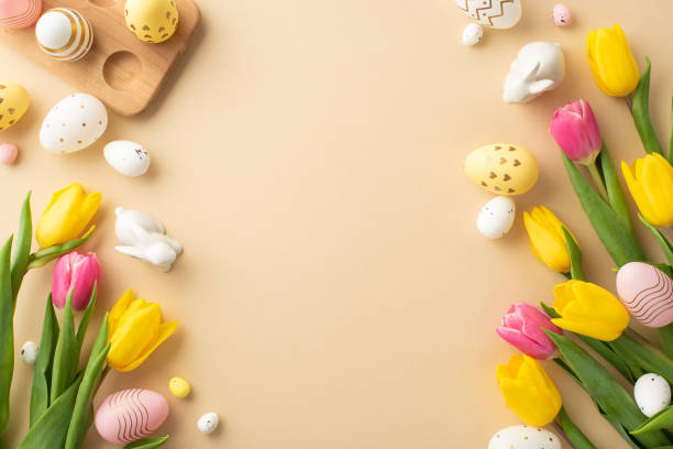 istock Easter decorations concept. Top view photo of colorful easter eggs ceramic easter bunnies yellow and pink tulips and wooden egg holder on isolated pastel beige background with empty space 1469401604