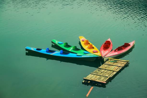 Colorful kayaks docked beside a white lighthouse on a calm urban river. Modern city skyline and construction cranes visible in the background.