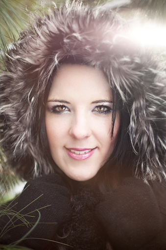 Wearing black coat with fluffy hood up, amongst green pine trees