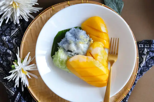 Top view of popular Thai dessert sweet Mango with sticky rice topping with coconut milk serve on white dish with golden fork put on handmade tray