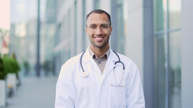 Portrait of an adult doctor in a white coat and glasses looking at the camera while standing on the street near a hospital building