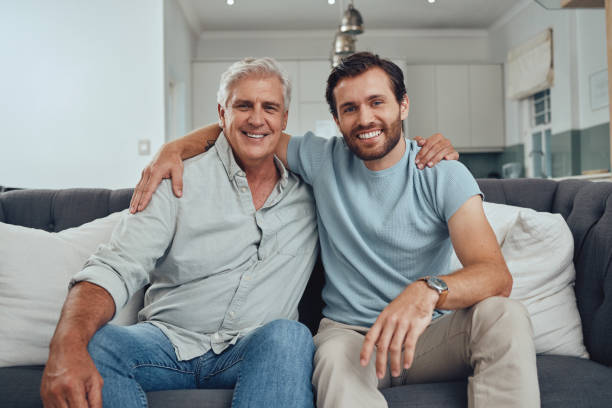 Portrait of a senior man with his adult son relaxing on a sofa together in the living room. Happy, smile and elderly male pensioner in retirement sitting and bonding with a young guy in family home. stock photo