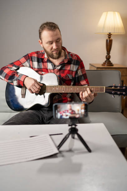istock Millennial man playing acoustic guitar while singing a song and record by cell phone for online social media. guitar class online live streaming with smartphone with student. 1469457229