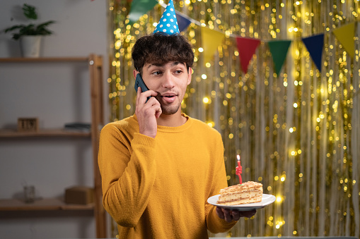 Young arab man celebrating birthday at home talking on cell phone holding birthday cake in hands. Copy space