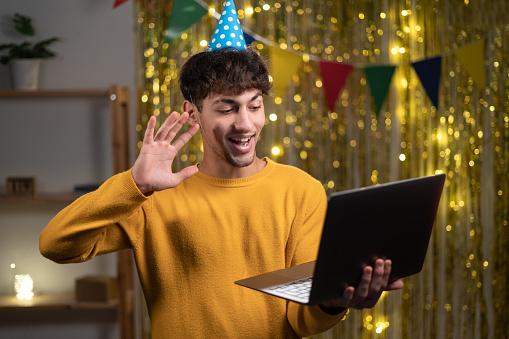 Virtual celebration concept. Portrait of happy young arabic man wearing party hat holding laptop and waving to webcam at home interior, making online video call on computer, greeting with birthday