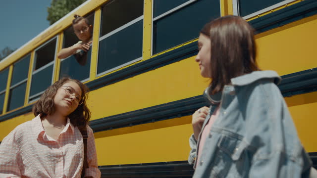 Two cheerful girls talking at school bus. Schoolboy looking out bus window.