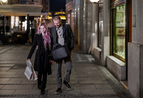 A happy young couple is enjoying the time on the city streets and has a nice conversation after successful shopping. A young woman and her husband are exchanging lovely emotions and enjoying a night in the city.