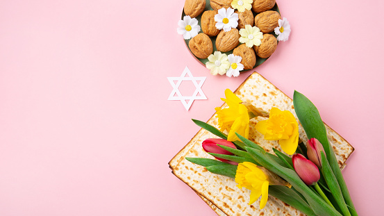 Jewish holiday Passover greeting card concept with matzah, Star of David, spring yellow daffodil, tulips, daisy flowers, walnuts on pink table. Seder Pesach spring holiday background, copy space.