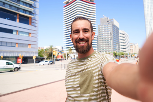 Copy space image of a happy caucasian man looking at camera while taking a selfie outdoors, standing against city road cheerfully. Point of view.