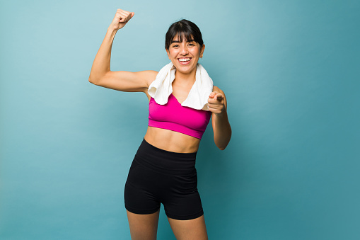 Cheerful strong woman with an athletic body showing her bicep muscle using a gym towel after her workout