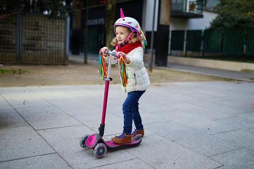 Adorable preschooler girl riding her scooter in a city park on sunny spring day. Cute child in unicorn helmet riding a push scooter. Active leisure and outdoor sport for kids