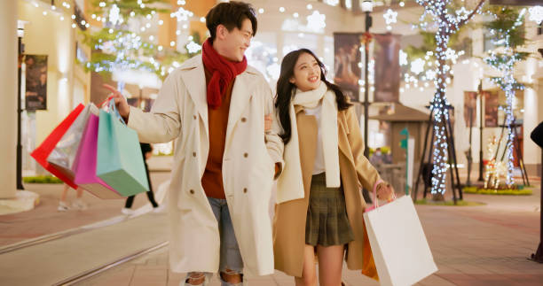 asian couple dating at night asian young couple wearing scarves with bags are chatting and walking on light decorated street while window shopping in winter at night window shopping at night stock pictures, royalty-free photos & images