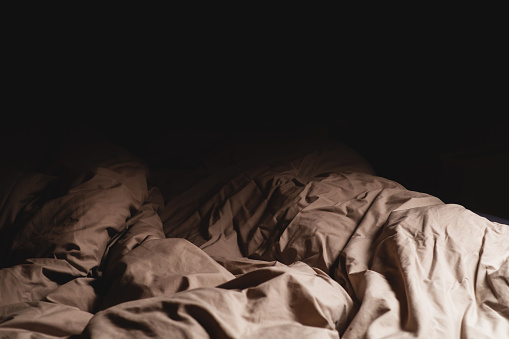 An empty unmade bed in the dark. A crumpled blanket lit by a lamp. Place for text.