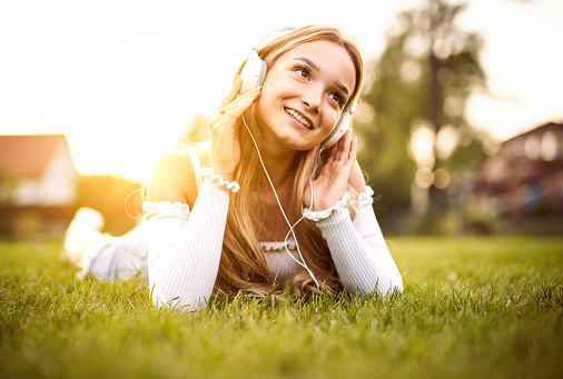 Teenager girl enjoying life and lying on front in the grass in a public park, while listening to music with headphones on her head with the sunset in the background on a sunny day