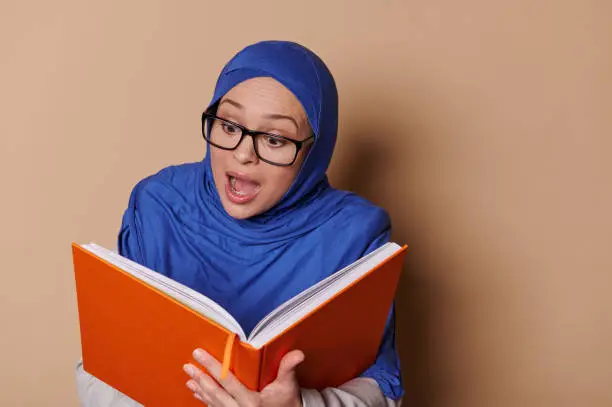 Amazed Arab Muslim woman wearing eyeglasses and blue hijab, expressing surprise and happy emotion while reading fairy tail, isolated on beige background. People. Education. Erudition. Learning concept