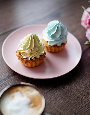 Two delicious, Sweet cream Cakes on a pink plate with yellow and blue cream sit on a wooden table. A cup of cappuccino with wooden Capcakes on the background. Close-up. Copy space