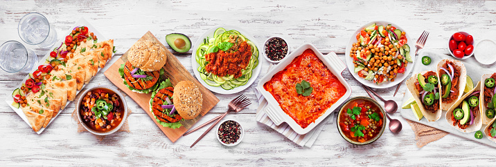 Healthy plant based vegetarian meal table scene. Overhead view on a white wood banner background. Jackfruit tacos, zucchini lasagna, walnut bolognese zoodles, chickpea burgers, hummus, soups, salad.