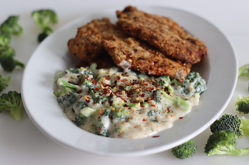 Breaded chicken served with sauteed broccoli in white sauce. Breaded chicken prepared in air fryer. Shot on white background with fresh broccoli around.