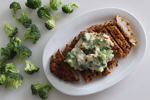 Breaded chicken served with sauteed broccoli in white sauce. Breaded chicken prepared in air fryer. Shot on white background with fresh broccoli around.