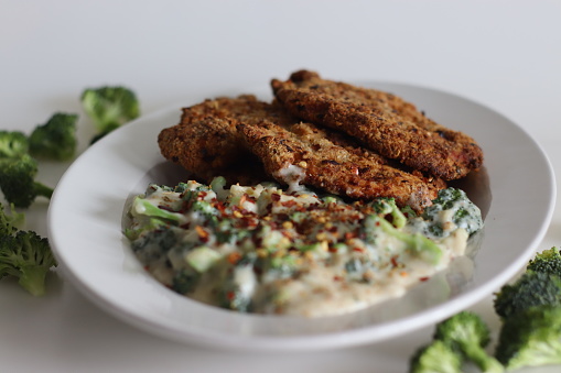 Breaded chicken served with sauteed broccoli in white sauce. Breaded chicken prepared in air fryer. Shot on white background with fresh broccoli around.