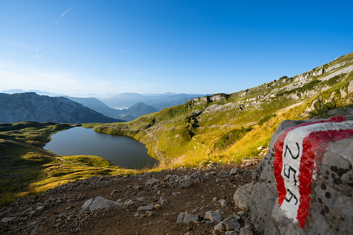 Beautiful summer morning high up in austrian alps with dark water in mountain lake and hiking path marker on stone, blue sky, sunny day