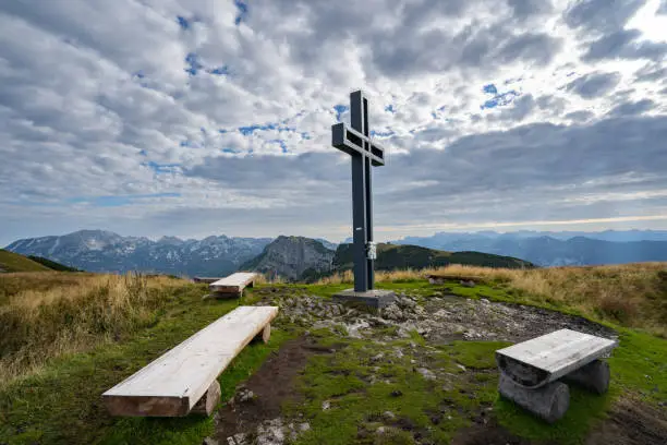Mountain peak with summit cross, wooden benches and cloudscape without people on summer morning in european alps in austria