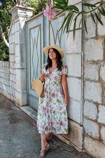 Portrait of beautiful girl wearing floral pattern dress and hat, posing under oleander flower in front of wall with gate