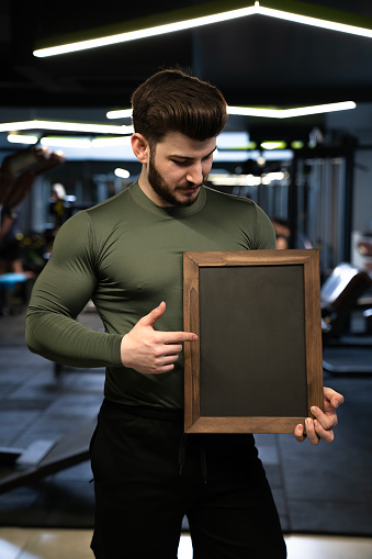A profile view of a powerful young adult man at the fitness center. Handsome young sporty sexy man showing a blackboard. Blank blackboard.