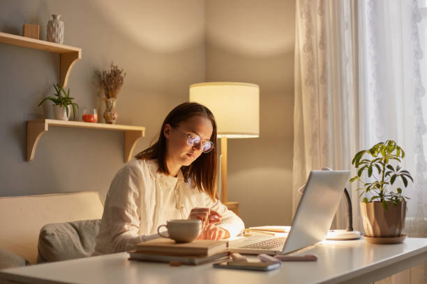 portrait d’une femme sérieuse et concentrée aux cheveux bruns assise à table travaillant sur un ordinateur portable le soir à la maison, écrivant et travaillant en ligne, passant du temps à apprendre, - women computer home interior brown hair photos et images de collection