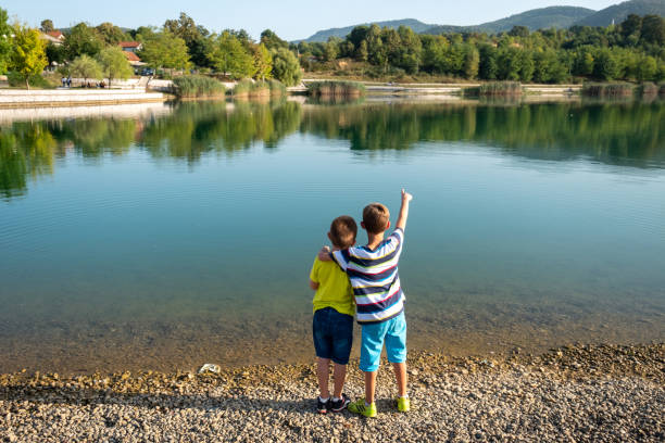 the boys have turned their backs to the camera while looking at the river and one boy is pointing something to the other by the river