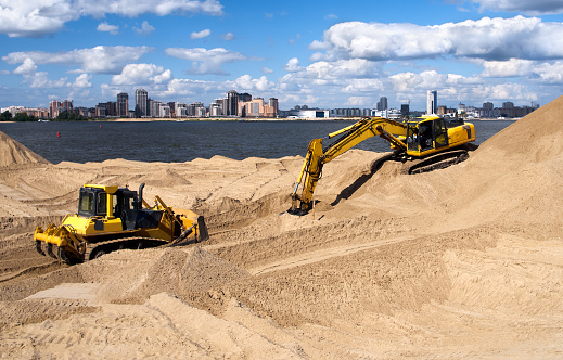Excavator on the bank of the river in the city digs the yellow sand of the beach, cloudy blue sky