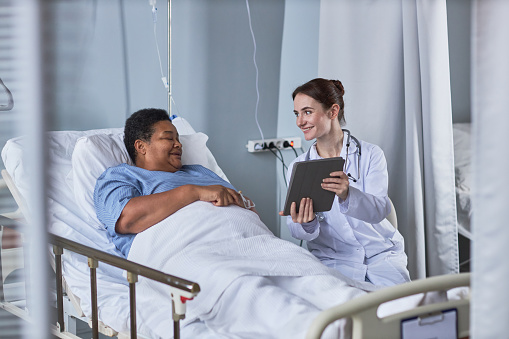 Portrait of smiling young nurse showing digital tablet to senior woman in hospital room