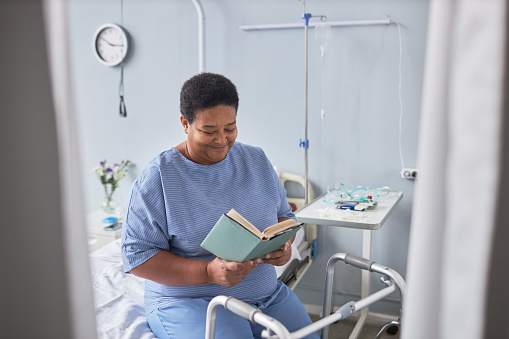 Portrait of smiling senior woman reading book in hospital room while sitting on bed in recovery